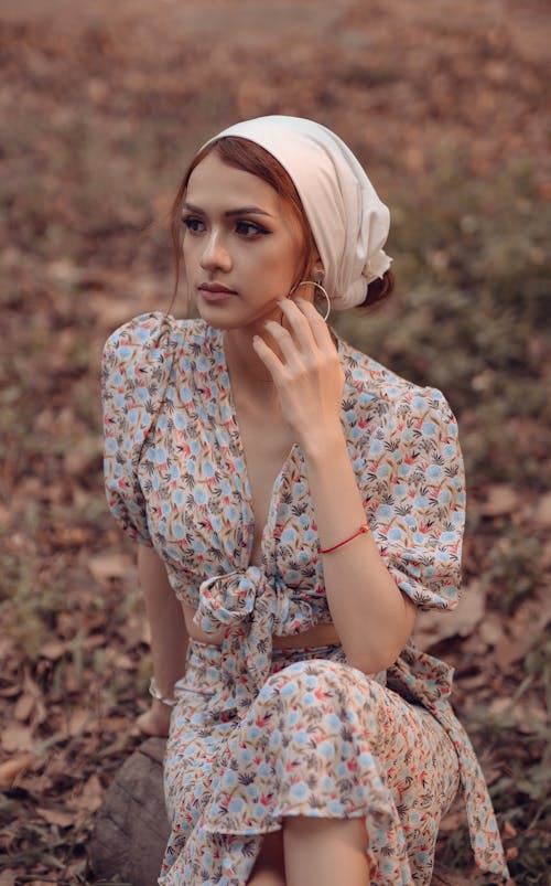 High angle of young thoughtful female in summer outfit touching cheek while sitting in countryside and looking away