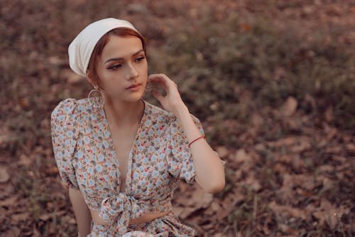 Free Young contemplative female model in summer dress and kerchief touching cheek while sitting on grass and looking away Stock Photo