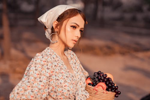 Charming female in summer outfit with fruits in wicker basket standing under sunlight while looking away