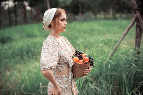 Side view of young female in summer clothes carrying basket full of ripe apples and grape while holding hand on hip and looking away