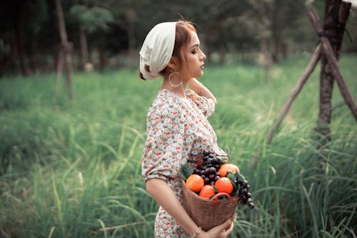 Side view of young female carrying basket with grape and apples while standing in countryside with closed eyes