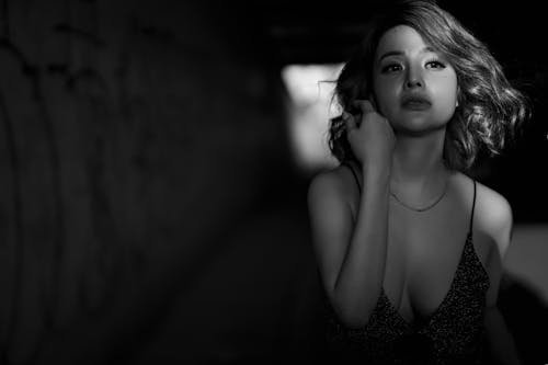 Black and white of slim ethnic female with wavy hair in elegant dress touching hair while looking away