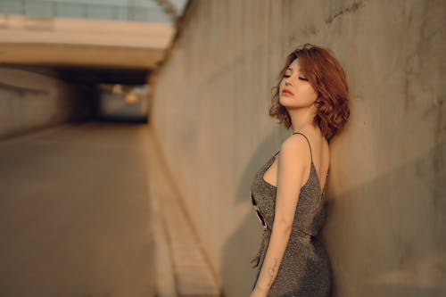 Side view of charming Asian female in trendy outfit leaning on wall with closed eyes