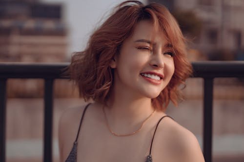 Young cheerful ethnic female with trendy hairstyle standing near railings while squinting and looking away