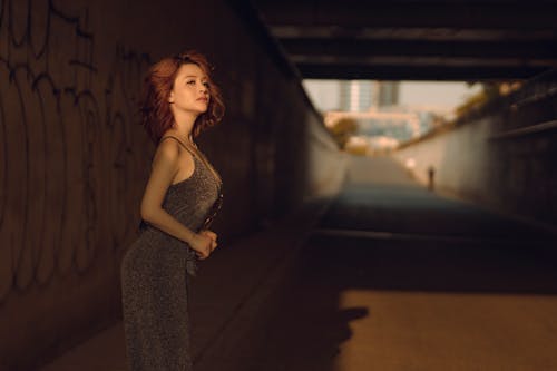 Side view of slim Asian female in stylish dress standing near wall with graffiti under sunlight and looking away
