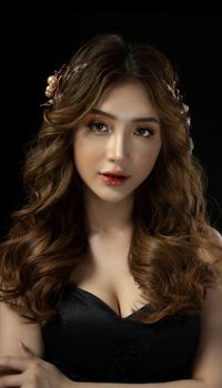 Young Asian woman in black clothes and makeup with wavy hairstyle and accessory on head in studio on black background and looking at camera