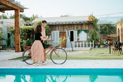 Free Full body side view of smiling young couple strolling on street with bicycle near pool and buildings near grass and plants in sunny summer day and looking at each other Stock Photo
