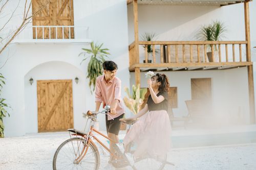Free Couple spending time in street near building with white wall and wooden doors with balcony while man riding woman with flower on bicycle near plants and looking at each other Stock Photo