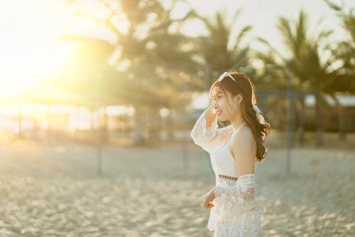 Side view of glad Asian female standing with hand on head standing on sandy coast with palms at sunset