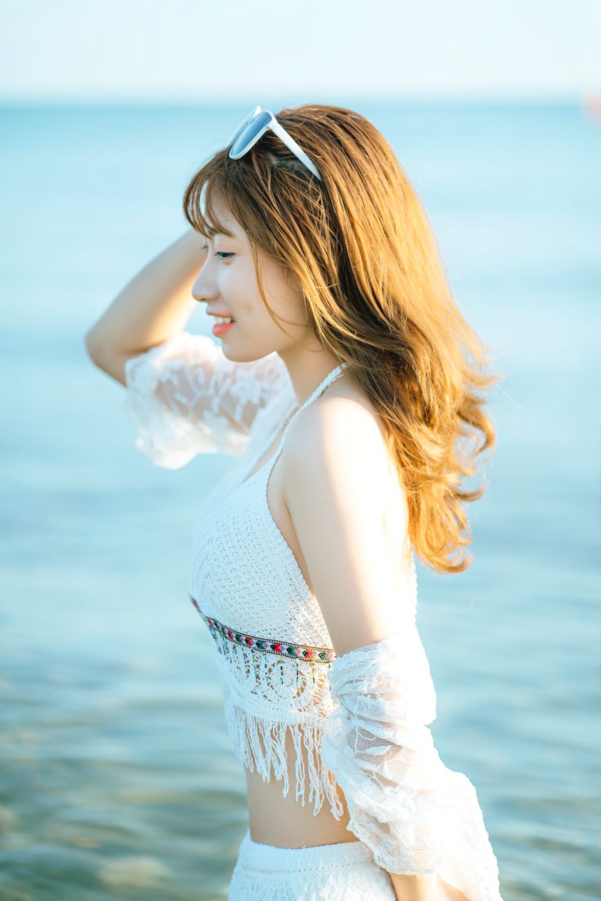Side view of smiling ethnic female traveler wearing white beachwear standing in seawater during trip