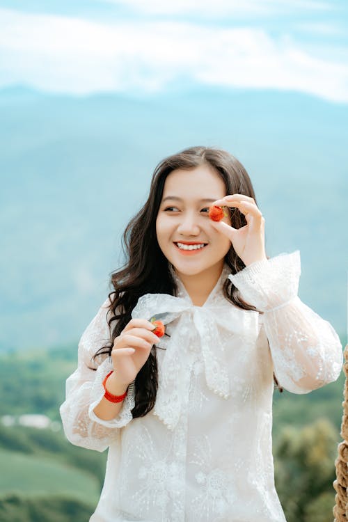 Free Young content Asian female in white apparel covering eye with ripe berry while looking away against mountain Stock Photo