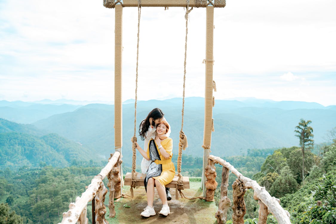 Asian daughter kissing content mother on swing against foggy ridges ...
