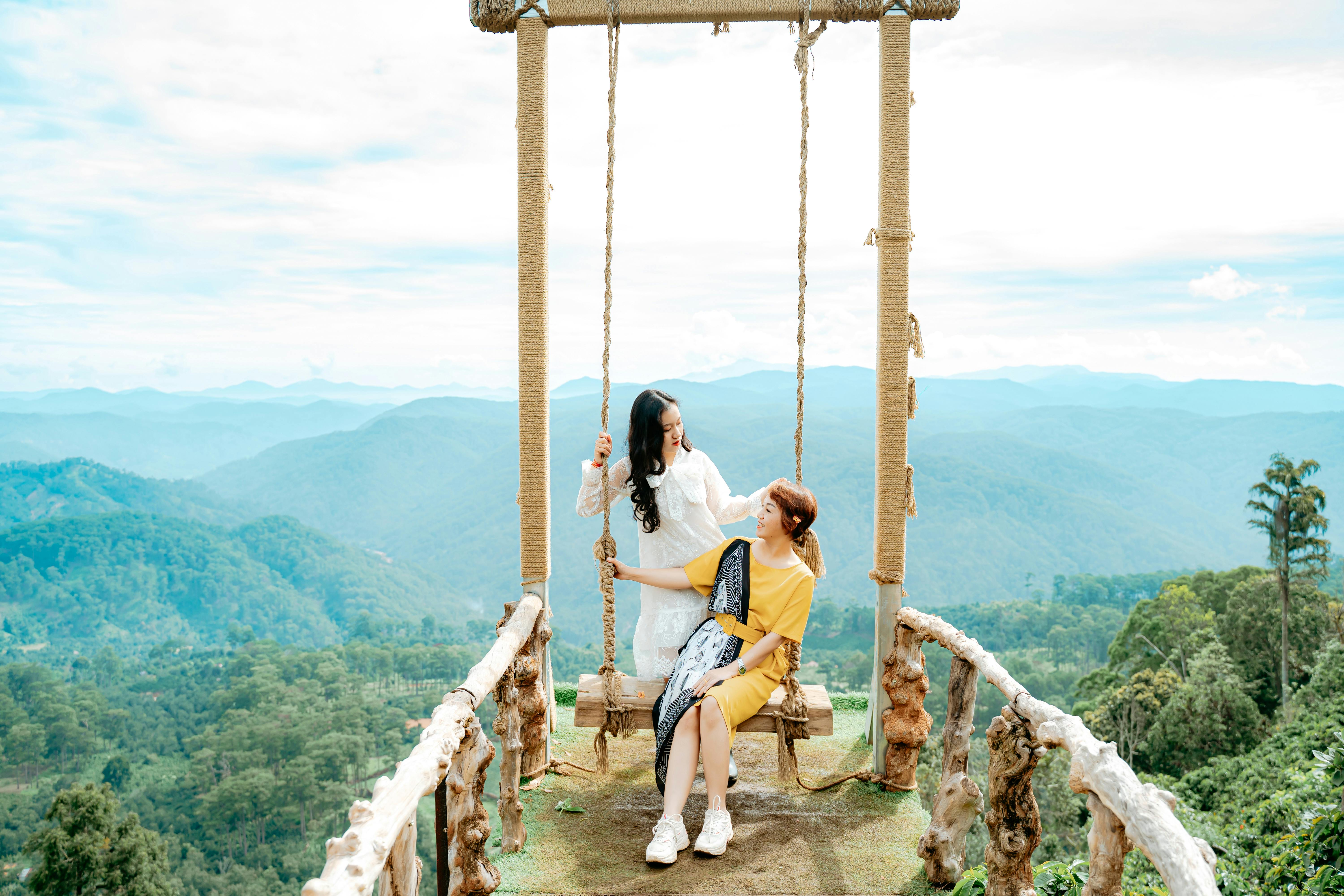 smiling ethnic mother with daughter talking on swing against ridges