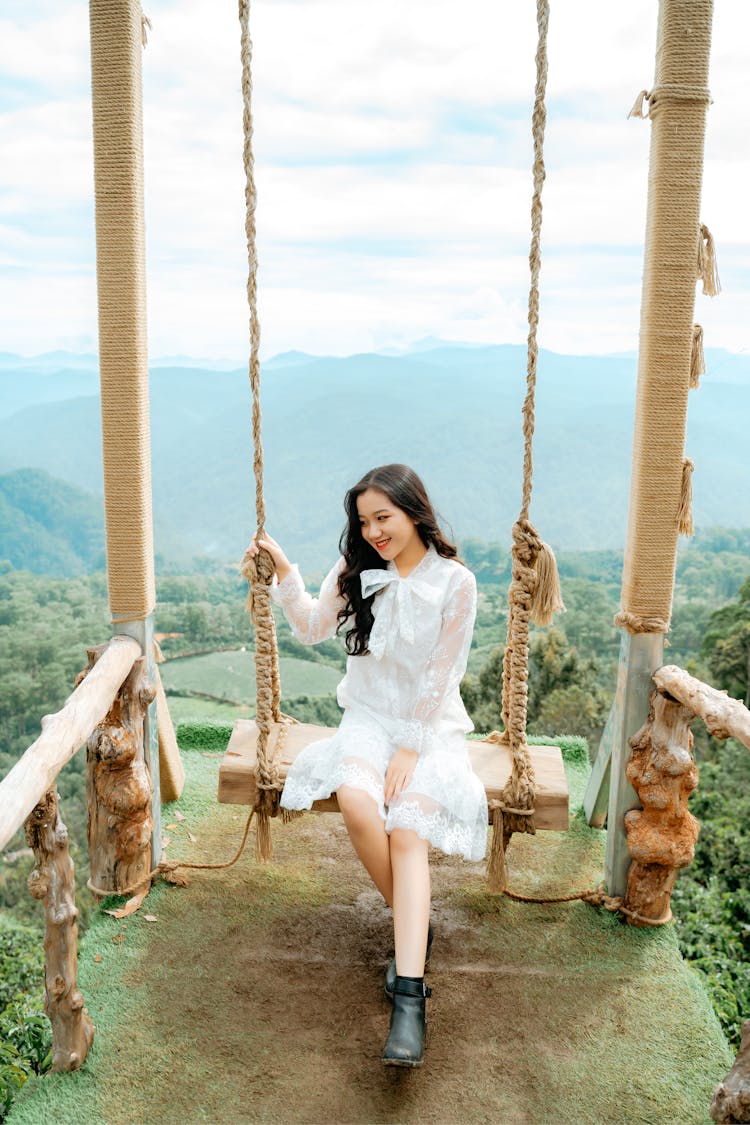 Cheerful Brunette Sitting On Swings On Grassy Hilltop