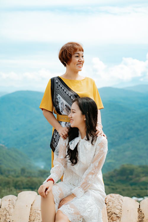 Cheerful young Asian female traveler in trendy dress smiling while resting in green mountainous valley with positive mother