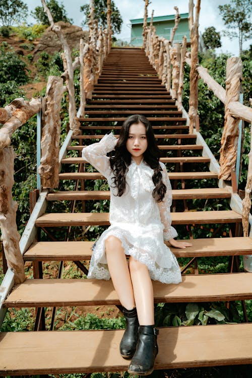 Full length of self assured young ethnic lady in stylish dress touching long dark hair and looking at camera while sitting on wooden staircase in countryside