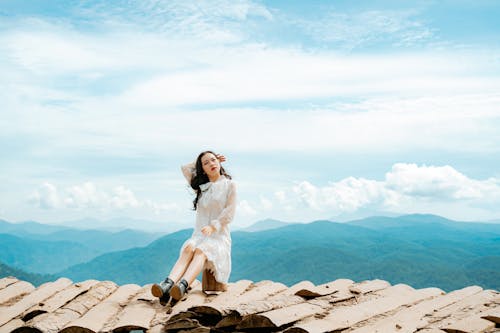 Full body of pensive young Asian female tourist in stylish dress touching long wavy hair and looking away while resting on shabby house rooftop against amazing mountains