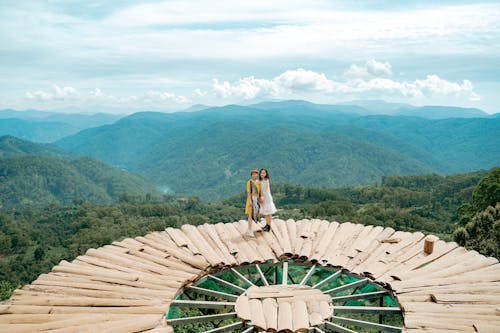 Side view of unrecognizable female travelers in stylish dresses standing together on creative wooden observation platform located in breathtaking green mountain valley