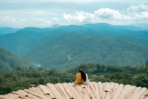 Back view of unrecognizable travelers resting on rough observation deck and admiring spectacular view of massive mountains covered with lush green trees