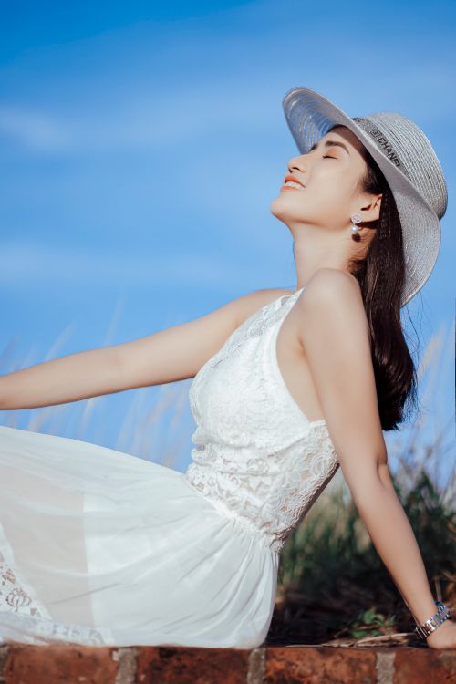 Free Low angle side view of relaxed smiling Asian female with closed eyes wearing straw hat and elegant white dress sitting on brick surface Stock Photo