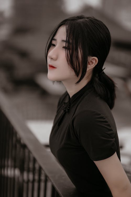High angle of young dreamy Asian female in dark clothes standing on street leaning on railing and looking away