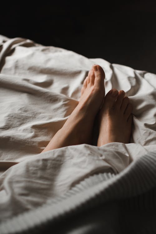 Free Crop anonymous barefoot female feet relaxing on soft comfy bed under white warm blanket in dark bedroom Stock Photo