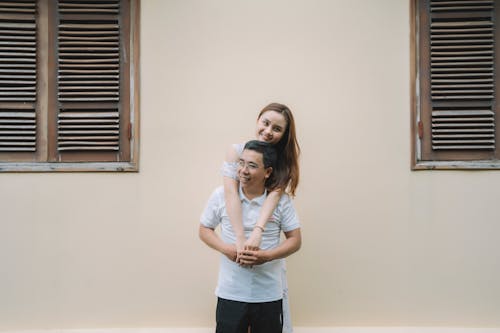 Cheerful young Asian man giving piggyback to girlfriend while enjoying sunny day on street together