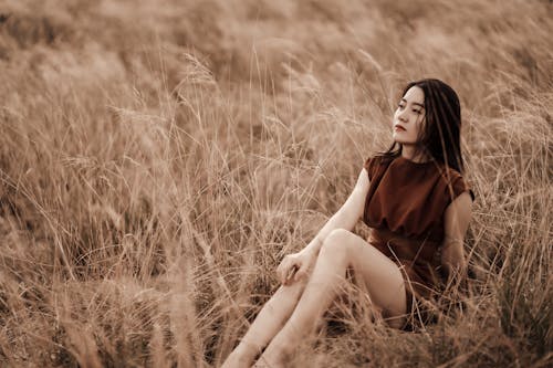 Young dreamy ethnic female in stylish apparel sitting on faded grass and looking up in daylight