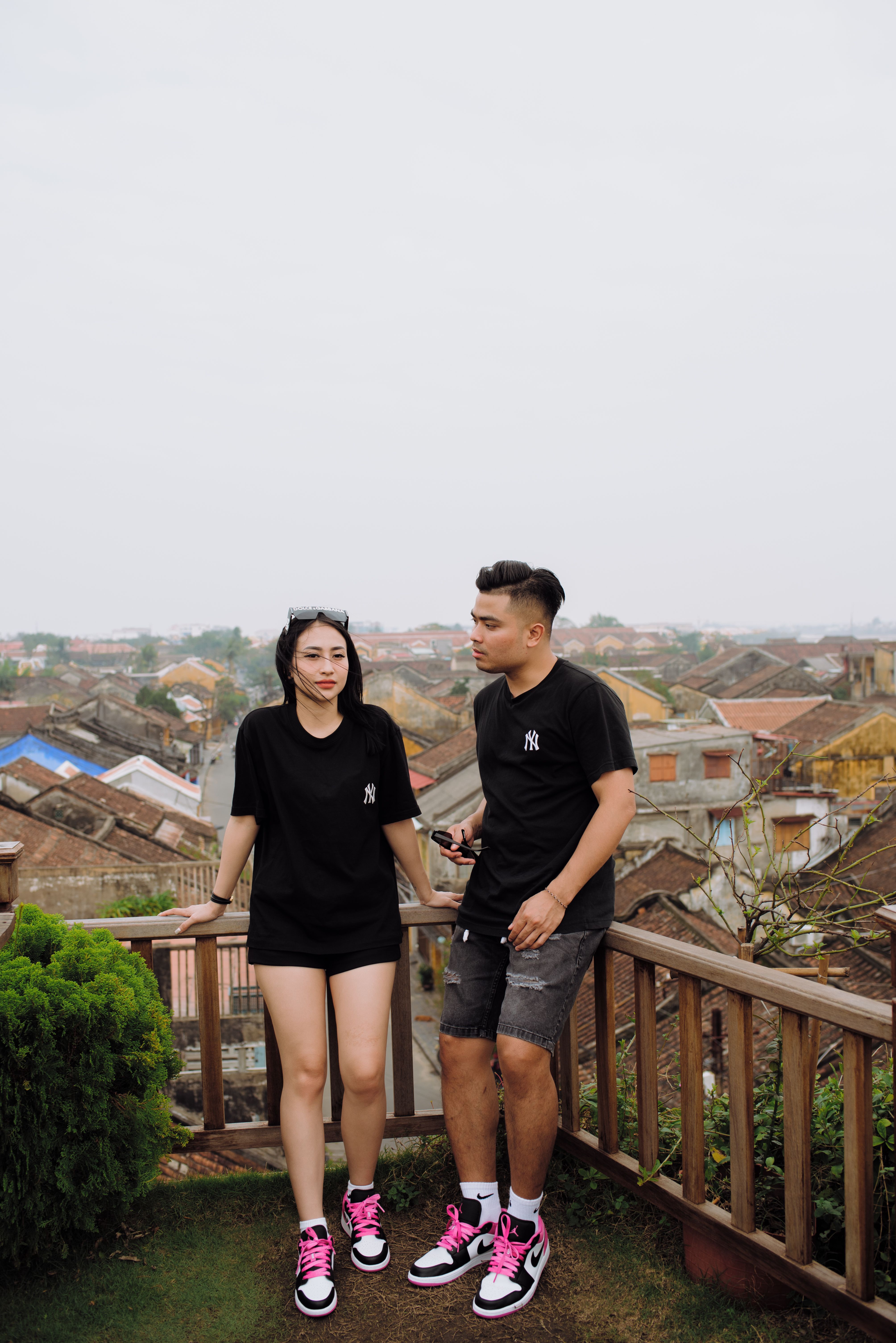 Young ethnic couple of tourists in modern sneakers standing on fenced veranda against aged urban houses under light sky