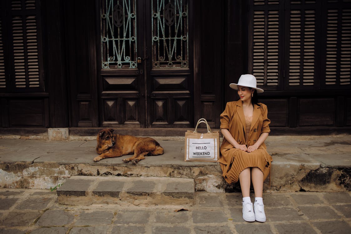 Free Young content ethnic female tourist in stylish apparel looking away near street dog while sitting on pavement in town Stock Photo