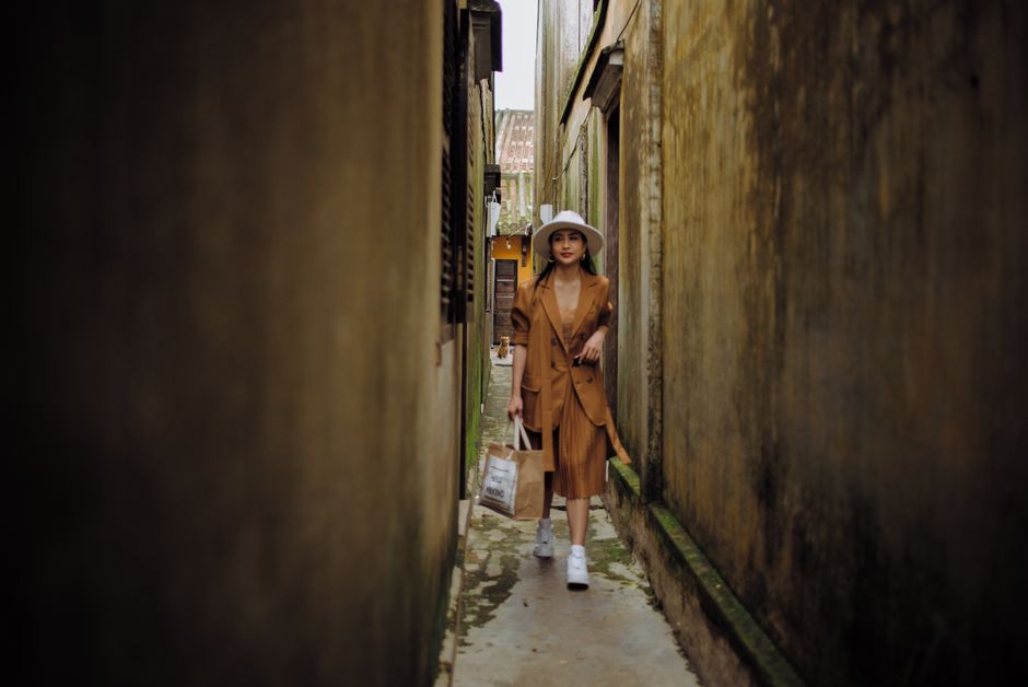 Young feminine ethnic female traveler walking on narrow pavement between weathered walls while looking away