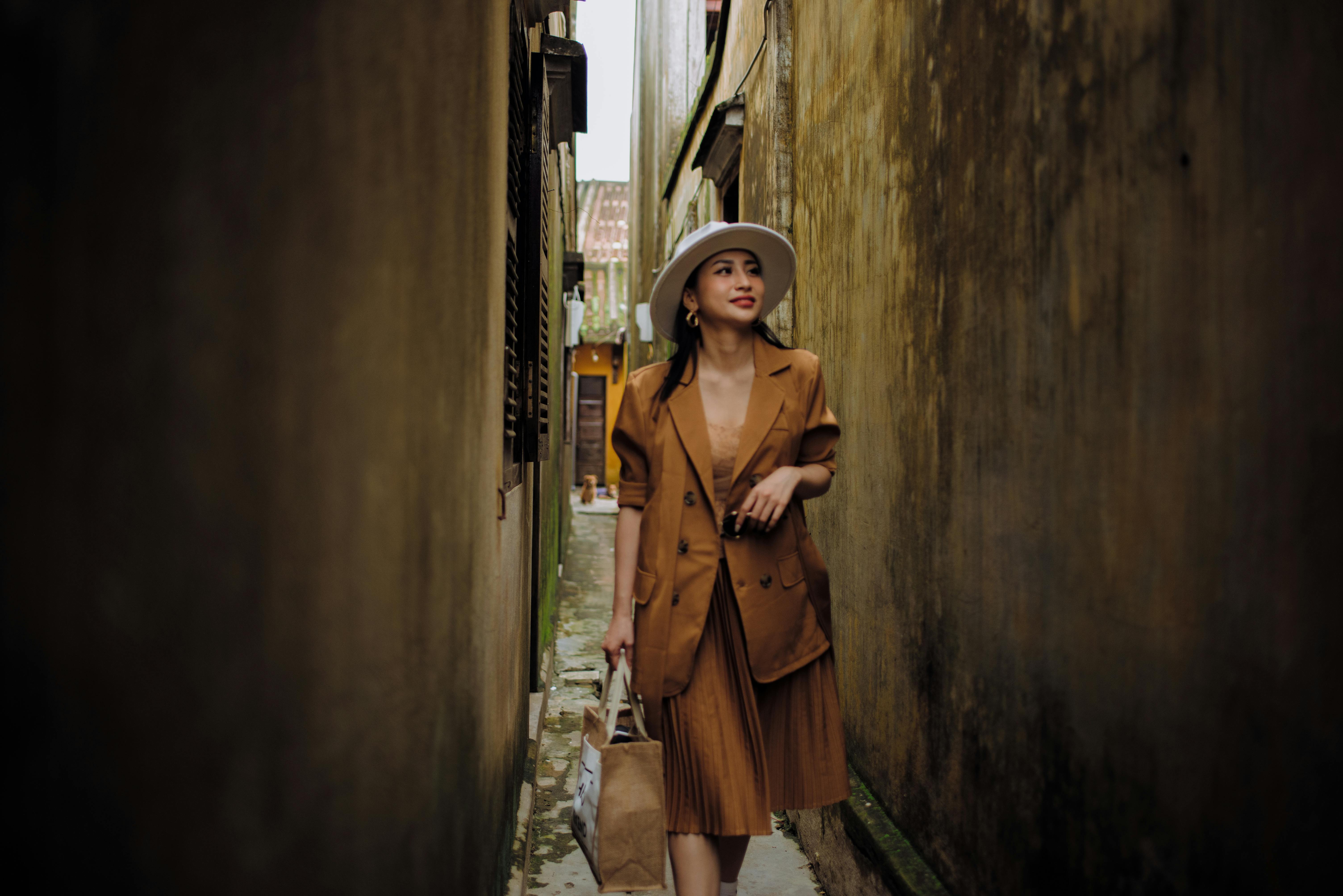 Young content ethnic female in trendy outfit strolling on narrow urban street while looking away