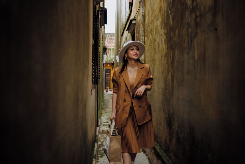 Young content ethnic female in trendy outfit strolling on narrow urban street while looking away