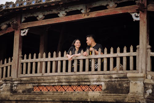 Happy young Asian couple leaning on wooden balcony railing while having pleasant conversation and looking at each other with smiles
