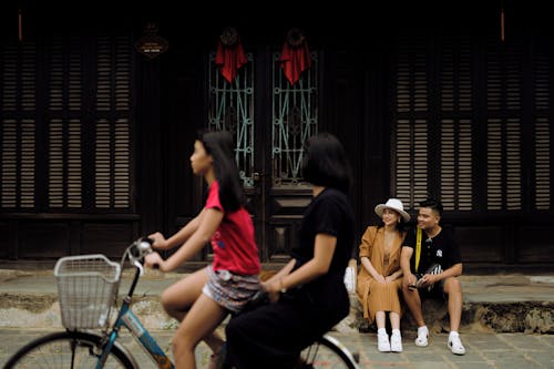 Cheerful Asian couple sitting on doorsteps near women riding bicycle