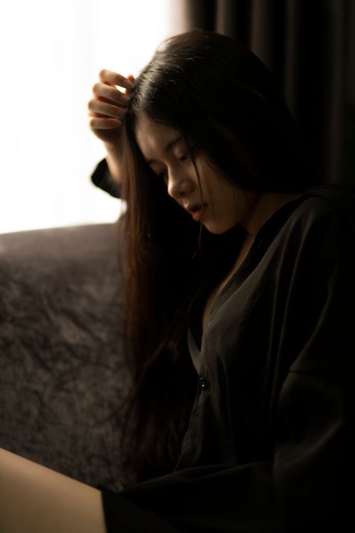 Free Sensitive young Asian female in black mini dress touching hair tenderly and sitting with eyes closed on comfy sofa Stock Photo