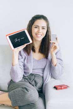 Young woman sitting on a couch holding a tablet and credit card, ready to shop online.