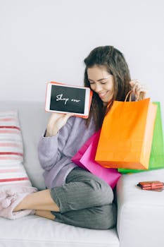 Woman holding a tablet and colorful shopping bags on a sofa, enjoying online shopping at home.
