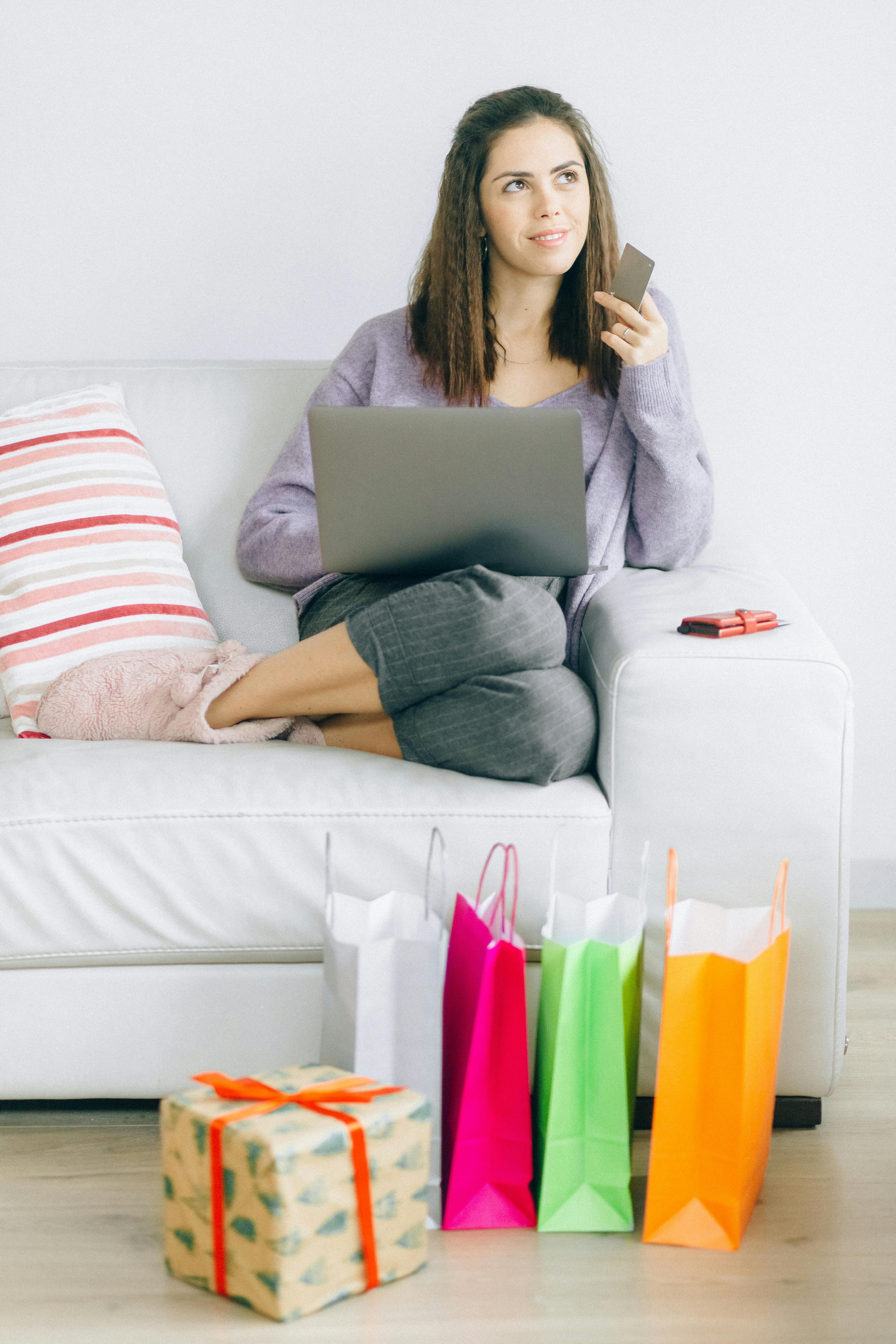 A woman sits comfortably on a sofa, using a laptop for online shopping with shopping bags nearby.