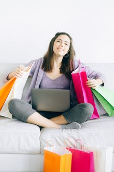 Smiling woman sits on a sofa with shopping bags and a laptop, embodying a joyful online shopping moment.