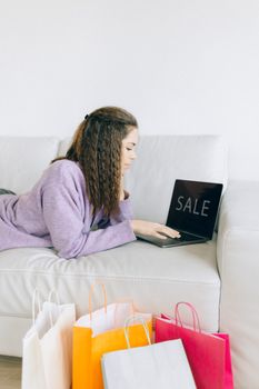 Woman browsing online sale while lying on a couch with shopping bags nearby.
