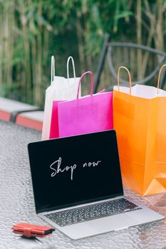Laptop displaying 'Shop now' with vibrant shopping bags on glass table outdoors.