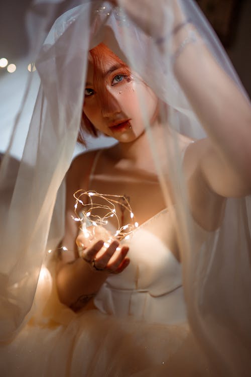 Young ethnic female in bridal dress with veil and shiny garland looking at camera in twilight