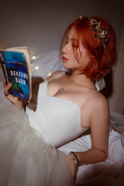 Young romantic Asian woman in white dress with adornment on head reading book in house