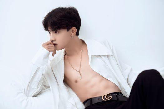 Charming Asian male with dark hair and naked torso in trendy outfit lying and thoughtfully looking away against white background