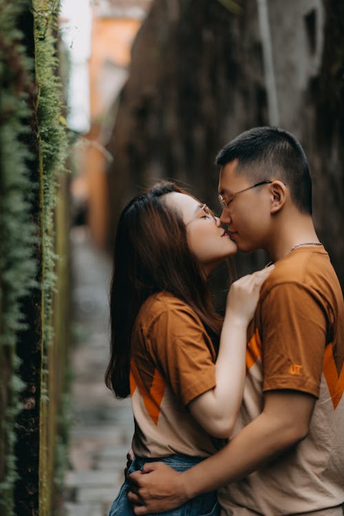 Side view of tender happy ethnic couple in eyeglasses kissing on urban street on blurred background of town