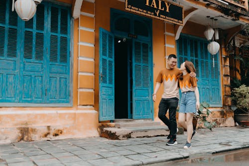 Full length of happy Asian couple in same t shirts hugging and looking at each other while walking on street near entrance of colorful building