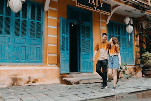 Free Full length of happy Asian couple in same t shirts hugging and looking at each other while walking on street near entrance of colorful building Stock Photo