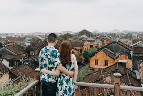 Back view of unrecognizable couple in similar wear hugging and looking away while standing on terrace of rooftop in gloomy day