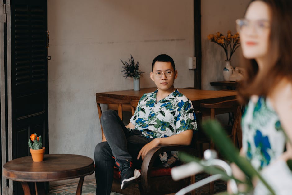 Focused Asian man in casual outfit and eyeglasses admiring satisfied Asian woman in same clothes and glasses while sitting in armchair in modern restaurant